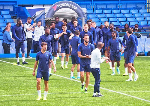 Elenco do Chelsea em treino no gramado do estádio, com jogadores de uniforme azul e as arquibancadas vazias ao fundo