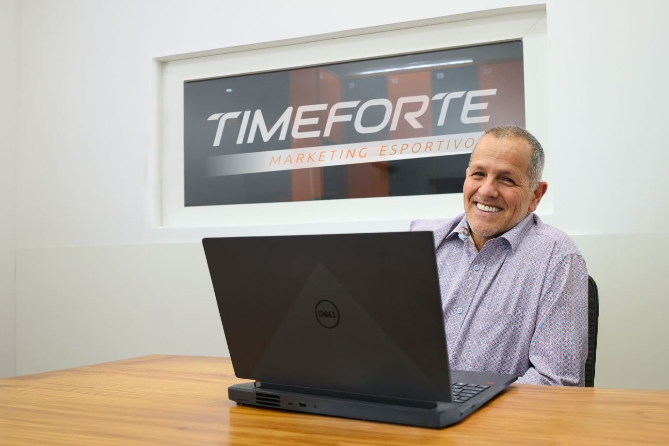 José Martins, fundador da Time Forte, sorri sentado à mesa com um notebook, diante do letreiro Time Forte Marketing Esportivo na parede do escritório