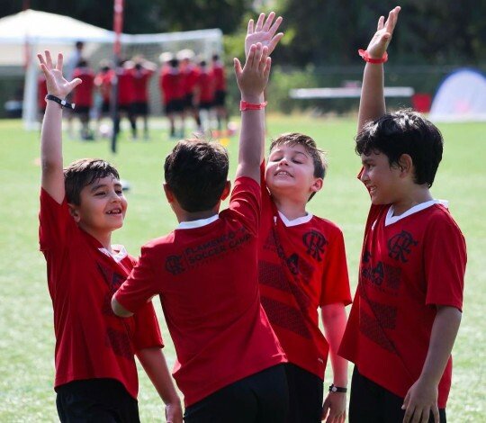 Quatro crianças com camisas vermelhas do Flamengo Soccer Camp comemoram de braços erguidos em um campo de futebol