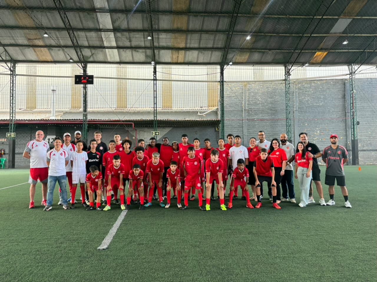Alunos de uniforme vermelho do Internacional posam com professores e dirigentes em quadra coberta de grama sintética da Escola do Inter