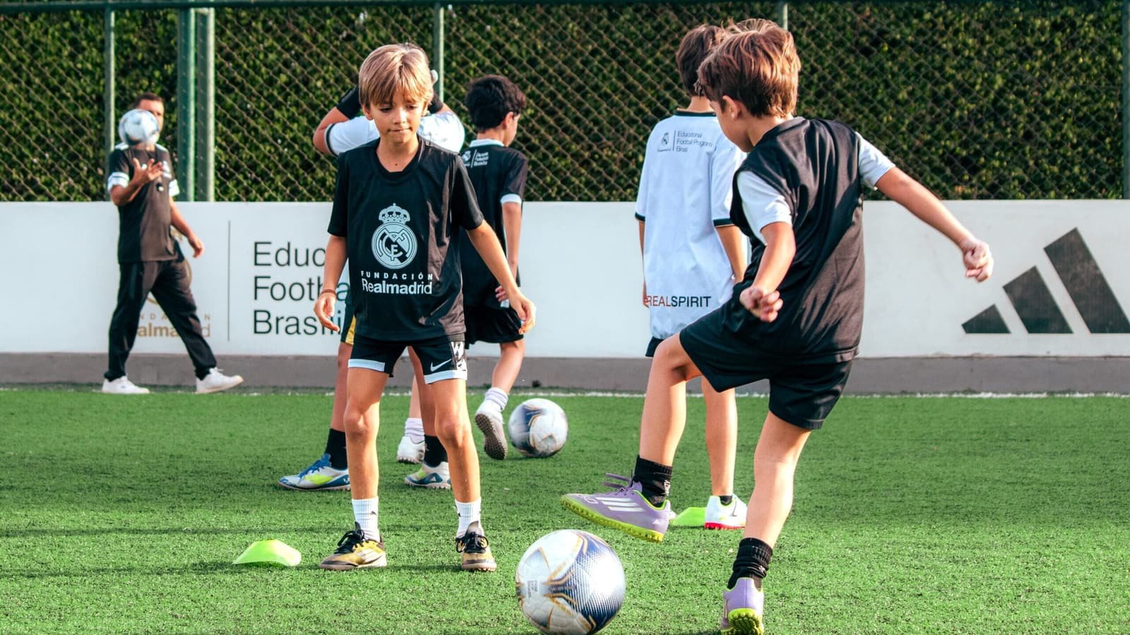Students at the Fundación Real Madrid school practising passes on an artificial pitch, wearing the foundation's black and white shirts, with the Educational Football Program Brasil board in the background