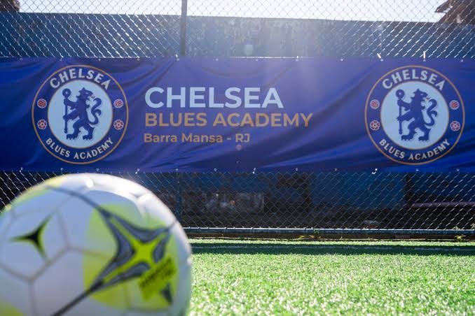 Banner azul da Chelsea Blues Academy com a inscrição "Barra Mansa - RJ" preso ao alambrado de um campo, com uma bola de futebol em primeiro plano