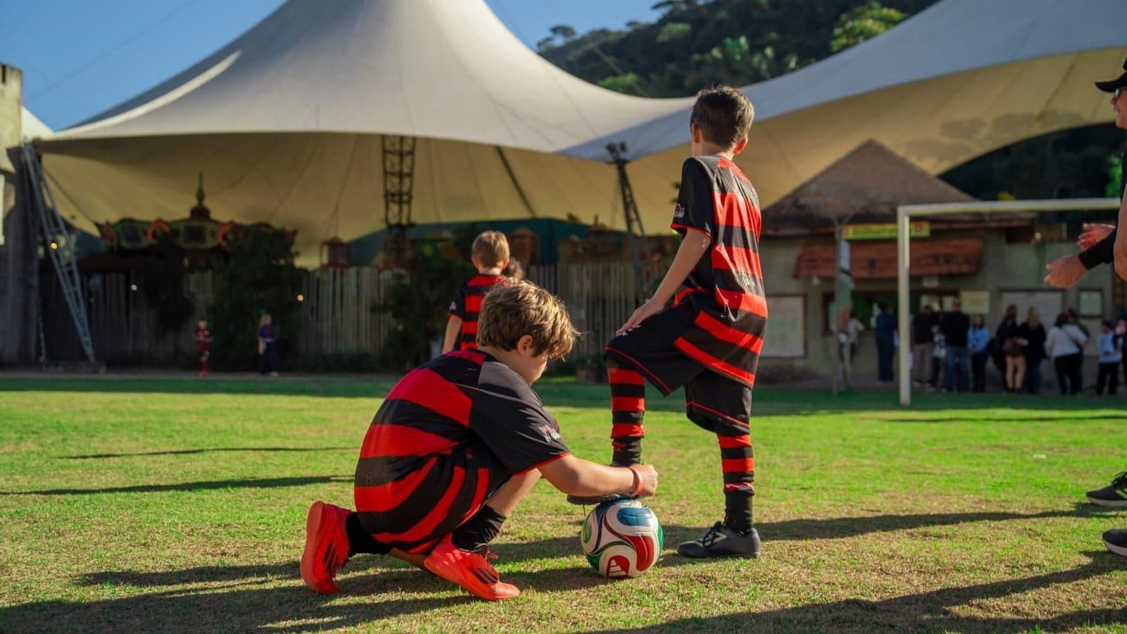 Two Escola Flamengo students in red-and-black kits on the grass — one crouching holds the ball while the other rests his foot on it — with a large white tent and a hill in the background